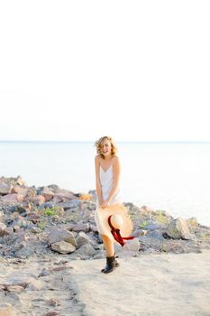 Caucasian blonde girl walking on rocky beach with hant in hands and wearing dress. Concept of summer vacations and fashion.