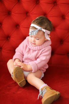 Little caucasian girl wearing pink sweater sitting on sofa with red background. Concept of baby photo session.