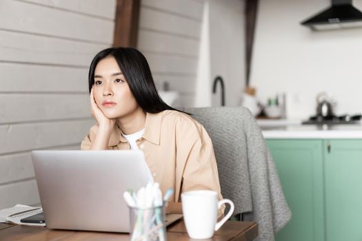 Bored asian girl student working on laptop, looking into distance with sad gloomy face, sitting at home, studying online.