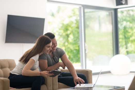 Young couple relaxing at luxurious home with tablet and laptop computers reading in the living room on the sofa couch.