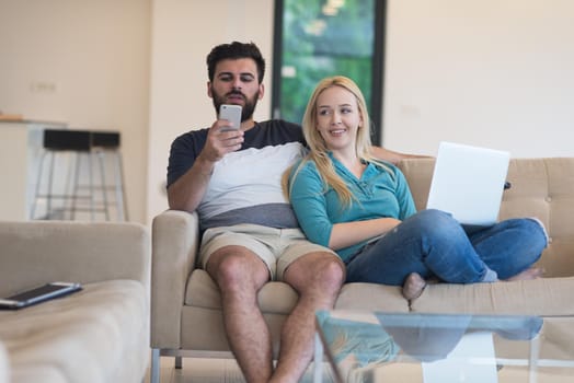 Young couple relaxes on the sofa in the luxury living room, using a laptop and remote control