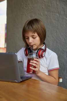 schoolgirl drinks lemonade through straw, uses laptop. little caucasian girl looking at computer, drinking red icy cocktail at table of cafe. modern communication technology, using digital devices