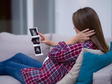 Young pregnant woman looking baby's ultrasound photo while relaxing on sofa at home