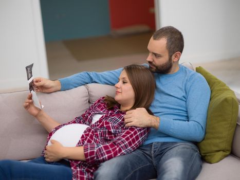 Young pregnant couple looking baby's ultrasound photo while relaxing on sofa at home