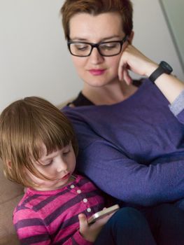 Beautiful young mother and her cute little daughter are using a mobile phone and smiling, sitting on sofa at home