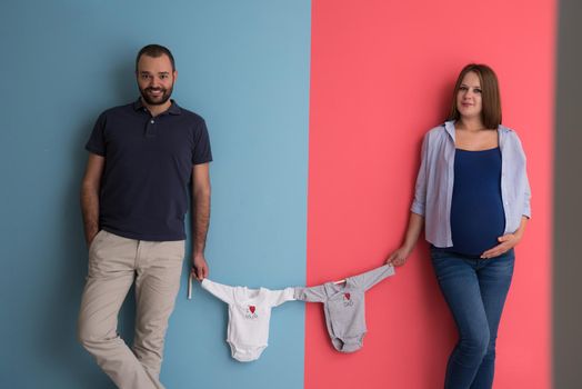 Beautiful pregnant woman and her husband expecting baby holding baby bodysuits and smiling over colorful background