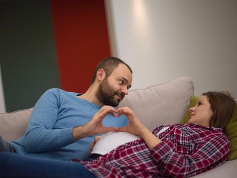 Happy man and pregnant woman showing heart sign with fingers while relaxing on the sofa at home