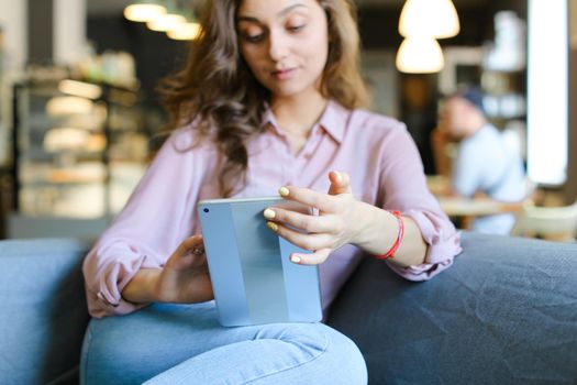 Young female happy student sitting on sofa at cafe and surfing by tablet. Concept of social networks, modern technology and free time with internet.