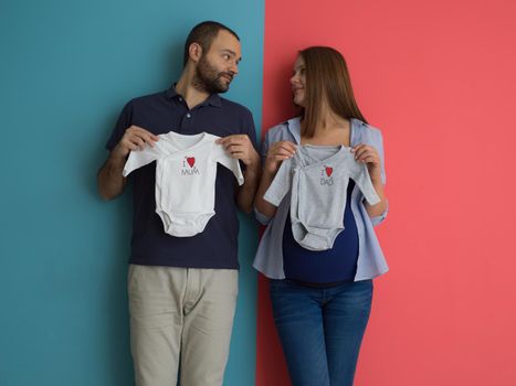 Beautiful pregnant woman and her husband expecting baby holding baby bodysuits and smiling over colorful background