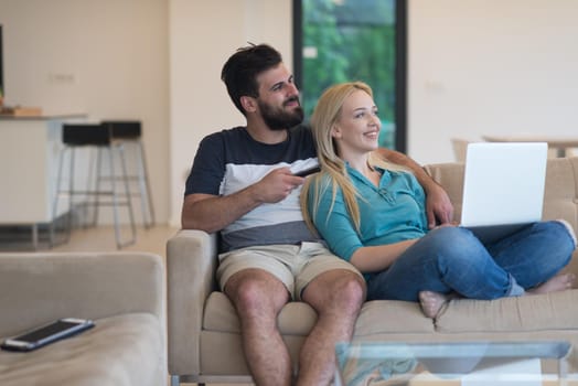 Young couple relaxes on the sofa in the luxury living room, using a laptop and remote control