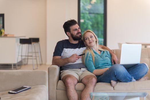 Young couple relaxes on the sofa in the luxury living room, using a laptop and remote control