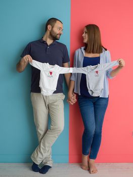 Beautiful pregnant woman and her husband expecting baby holding baby bodysuits and smiling over colorful background