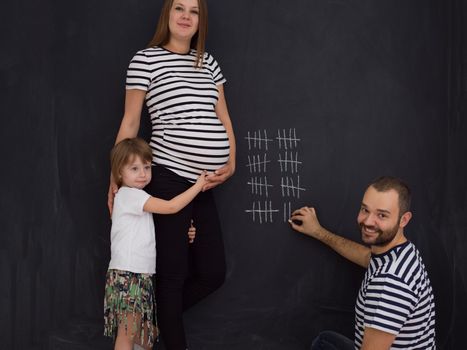 young pregnant couple with cute little daughter accounts week of pregnancy and writing them with chalk on blackboard