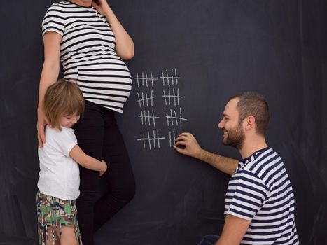 young pregnant couple with cute little daughter accounts week of pregnancy and writing them with chalk on blackboard