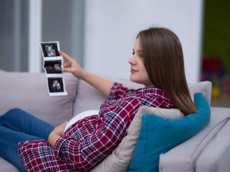 Young pregnant woman looking baby's ultrasound photo while relaxing on sofa at home