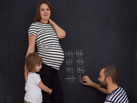 young pregnant couple with cute little daughter accounts week of pregnancy and writing them with chalk on blackboard