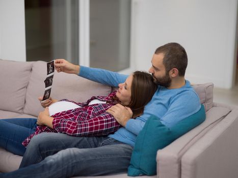 Young pregnant couple looking baby's ultrasound photo while relaxing on sofa at home