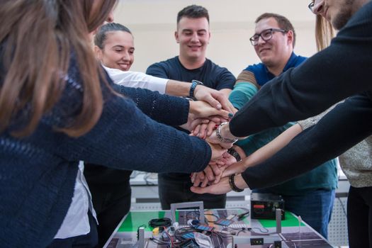 Group of young students in electronics classroom celebrating successfully finished project with holding their hands together, education and technology concept