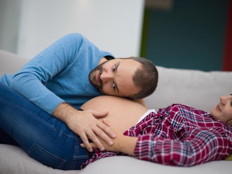 Happy future dad listening the belly of his pregnant wife while relaxing on sofa at home