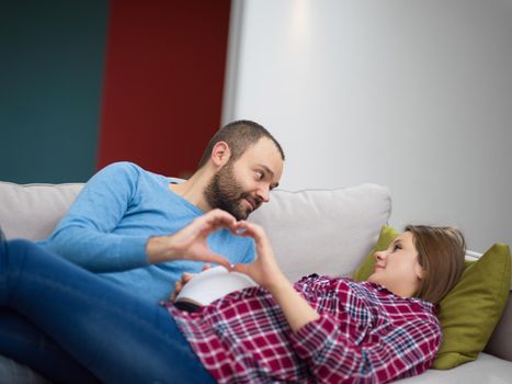 Happy man and pregnant woman showing heart sign with fingers while relaxing on the sofa at home