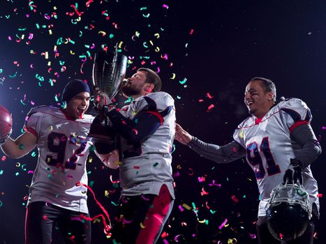 happy american football team celebrating victory with trophy and confetti on the night field