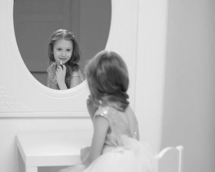 A nice little girl near the big mirror paints her lips with lipstick. The concept of childhood, style and fashion.