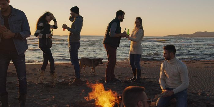 Happy Carefree Young Friends Having Fun And Drinking Beer By Bonefire On The Beach As The Sun Begins To Set