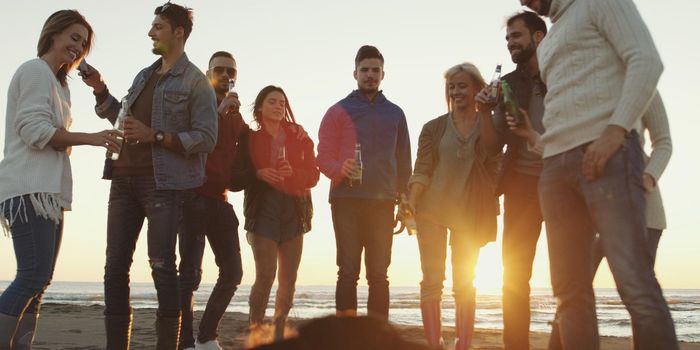 Happy Carefree Young Friends Having Fun And Drinking Beer By Bonefire On The Beach As The Sun Begins To Set