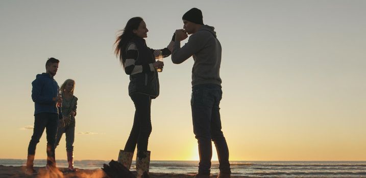 Happy Carefree Young Friends Having Fun And Drinking Beer By Bonefire On The Beach As The Sun Begins To Set