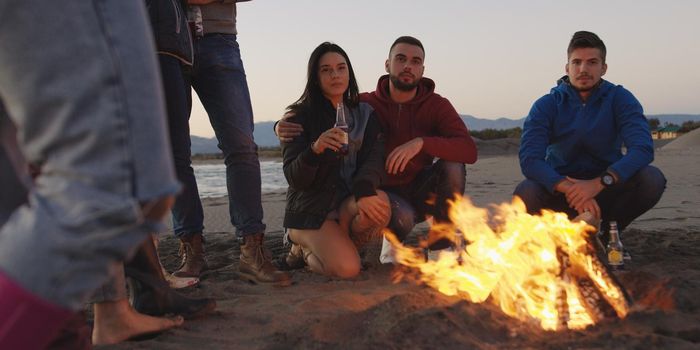 Happy Carefree Young Friends Having Fun And Drinking Beer By Bonefire On The Beach As The Sun Begins To Set