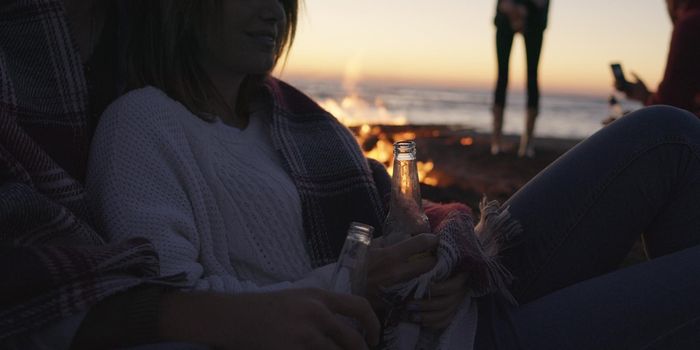 Happy Carefree Young Friends Having Fun And Drinking Beer By Bonefire On The Beach As The Sun Begins To Set