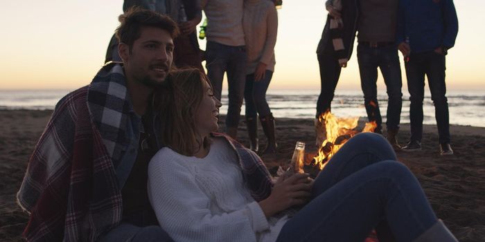 Happy Carefree Young Friends Having Fun And Drinking Beer By Bonefire On The Beach As The Sun Begins To Set