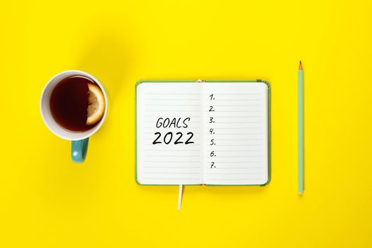 2022 goals banner with copy space. Notepad, pencil and cup of tea on a yellow background. Copy space, Flat lay. Women hand write goals for 2022 year