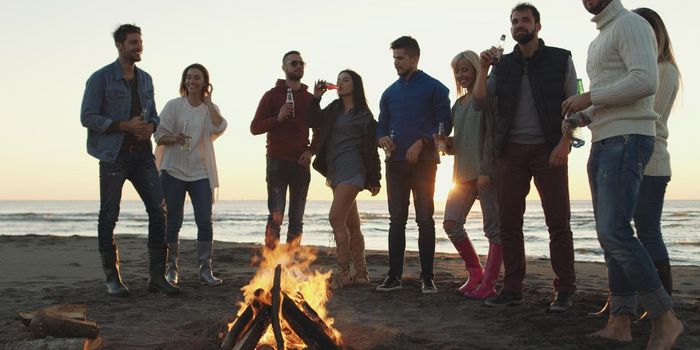 Happy Carefree Young Friends Having Fun And Drinking Beer By Bonefire On The Beach As The Sun Begins To Set