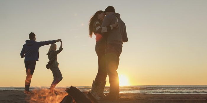 Happy Carefree Young Friends Having Fun And Drinking Beer By Bonefire On The Beach As The Sun Begins To Set