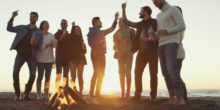 Happy Carefree Young Friends Having Fun And Drinking Beer By Bonefire On The Beach As The Sun Begins To Set