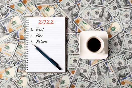 2022, GOAL, PLAN and ACTION. Notepad, pen and a cup of black coffee isolated on money background. Notepad with copy space. Office, business, discreet style.