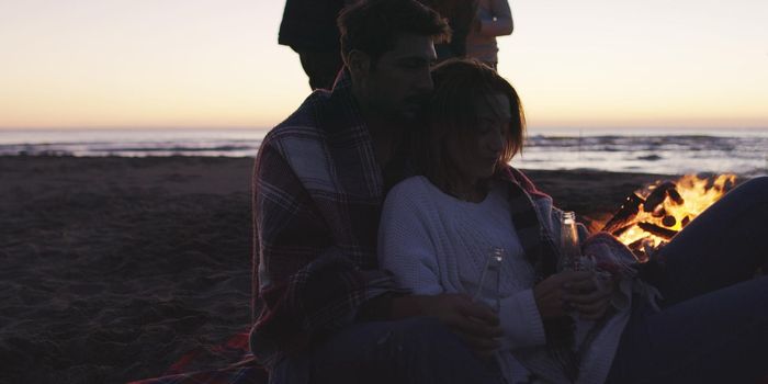 Happy Carefree Young Friends Having Fun And Drinking Beer By Bonefire On The Beach As The Sun Begins To Set