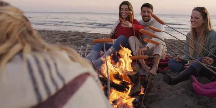 Group of young friends sitting by the fire late at night, grilling sausages and drinking beer, talking and having fun