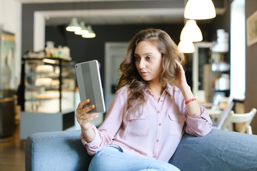 Young female student sitting on sofa at cafe and surfing by tablet. Concept of social networks, modern technology and free time with internet.