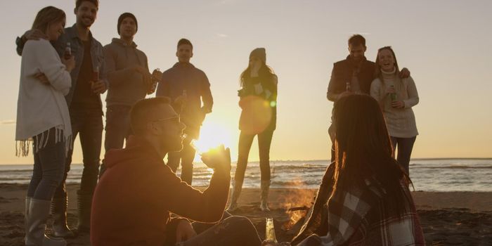 Happy Carefree Young Friends Having Fun And Drinking Beer By Bonefire On The Beach As The Sun Begins To Set