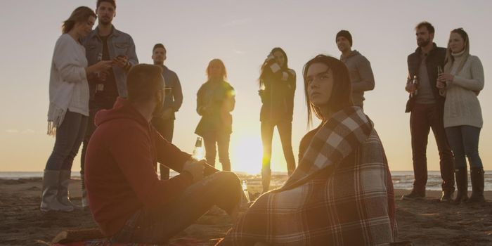 Happy Carefree Young Friends Having Fun And Drinking Beer By Bonefire On The Beach As The Sun Begins To Set