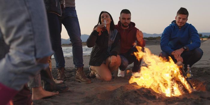 Happy Carefree Young Friends Having Fun And Drinking Beer By Bonefire On The Beach As The Sun Begins To Set