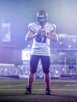 portrait of confident American football player holding ball while standing on the big modern stadium with lights and flares at night