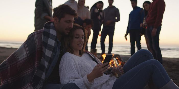 Couple using cell phone during beach party with friends drinking beer and having fun