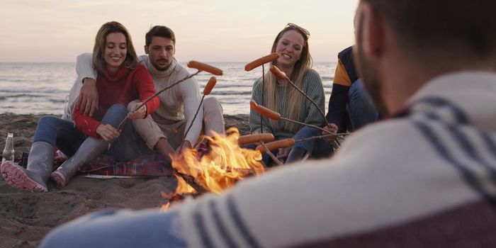 Group of young friends sitting by the fire late at night, grilling sausages and drinking beer, talking and having fun