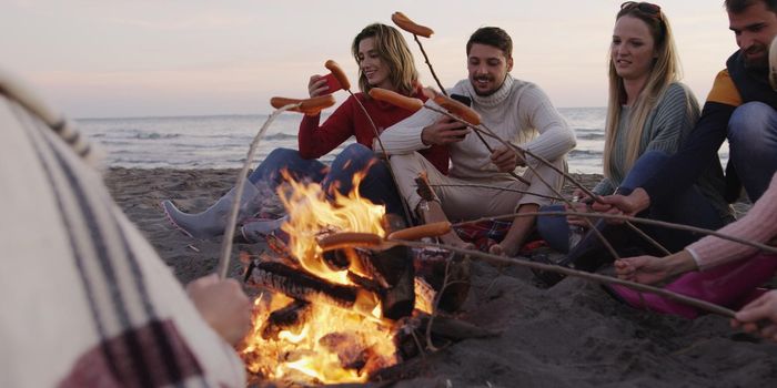 Group of young friends sitting by the fire late at night, grilling sausages and drinking beer, talking and having fun