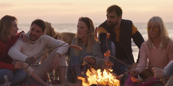 Group of young friends sitting by the fire late at night, grilling sausages and drinking beer, talking and having fun