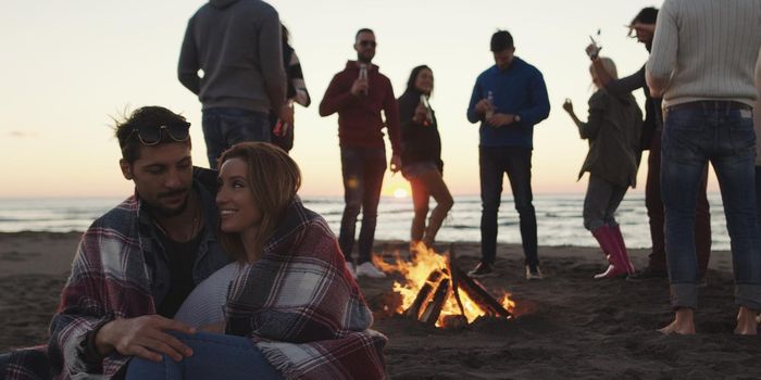 Happy Carefree Young Friends Having Fun And Drinking Beer By Bonefire On The Beach As The Sun Begins To Set