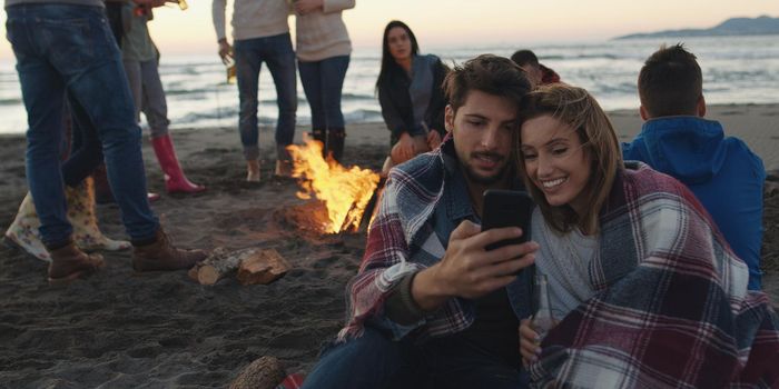 Couple using cell phone during beach party with friends drinking beer and having fun